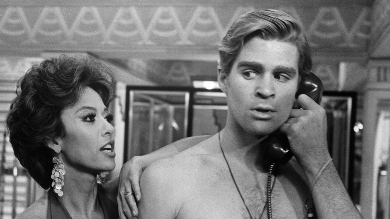 A shirtless Treat Williams holding phone next to Rita Moreno