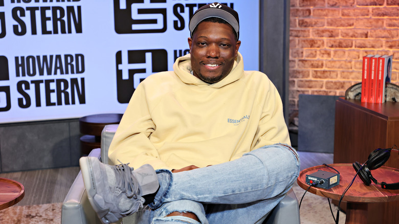 Michael Che seated with leg up