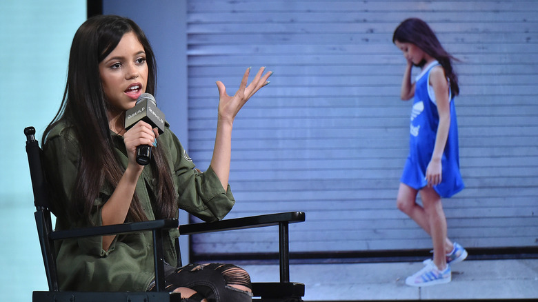 Jenna Ortega wearing an army green jacket and ripped black jeans talking into a microphone in front of a background of one of her characters on screen