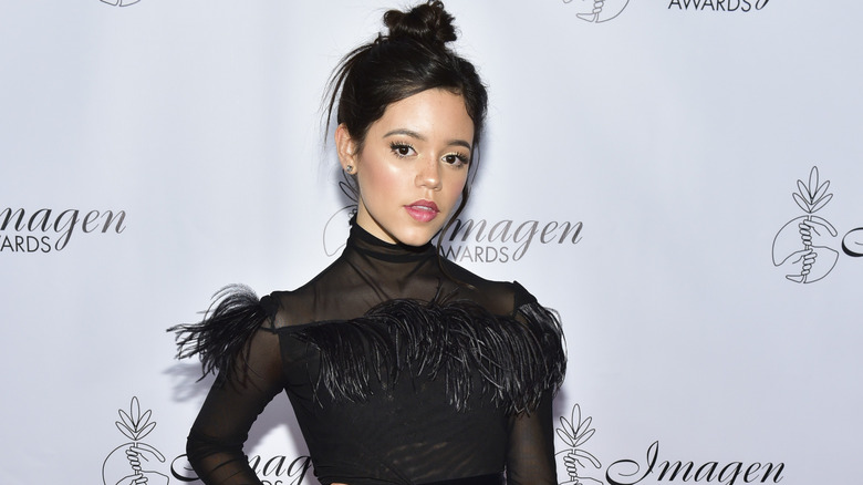 Jenna Ortega wearing a high neck black mesh dress with a feathered bust with her dark hair in a top knot bun