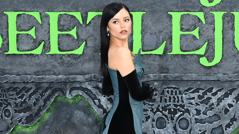 Jenna Ortega wearing a green and black gown with long back hair in front of a Beetlejuice Beetlejuice backdrop