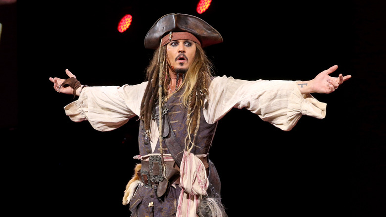 Johnny Depp wearing dreadlocked hair, billowing shirt, vest, sashes, and pirate hat