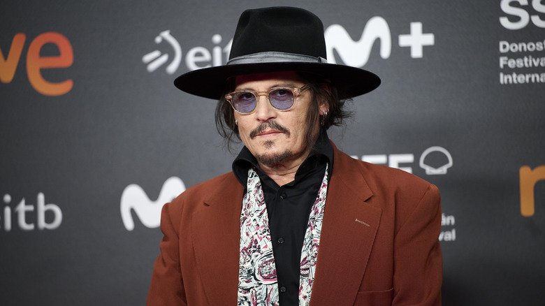 Johnny Depp wearing a burnt orange-colored jacket, paisley scarf, black shirt, black hat, and tinted sunglasses