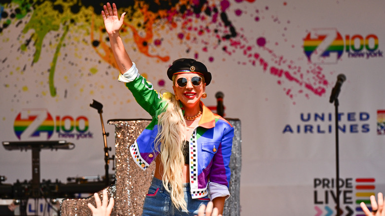 Lady Gaga smiling and holding her hand up wearing a rainbow jacket and black beret