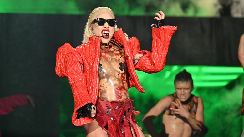 Lady Gaga performs wearing a red jacket, black sunglasses, and a see through red and flesh toned outfit