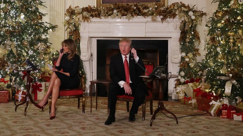 Melania and Donald Trump speaking on phones between Christmas trees at the White House