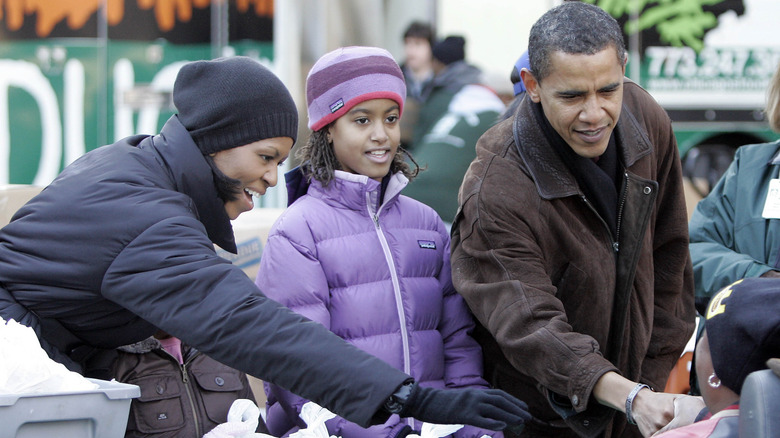 Michelle Obama wearing a blue winter coat and black beanie with Malia Obama wearing a purple winter coat and matching hat with Barack Obama wearing a brown coat