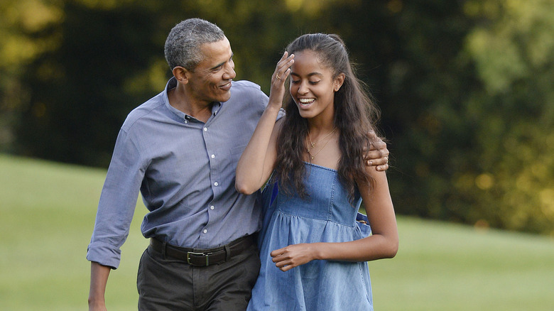 Barack Obama wearing a grey button down shirt and brown slacks with Malia Obama wearing a strappy denim sundress