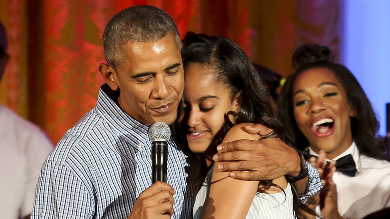 Barack Obama wearing a striped button down shirt hugging his daughter Malia, wearing a striped strapless dress