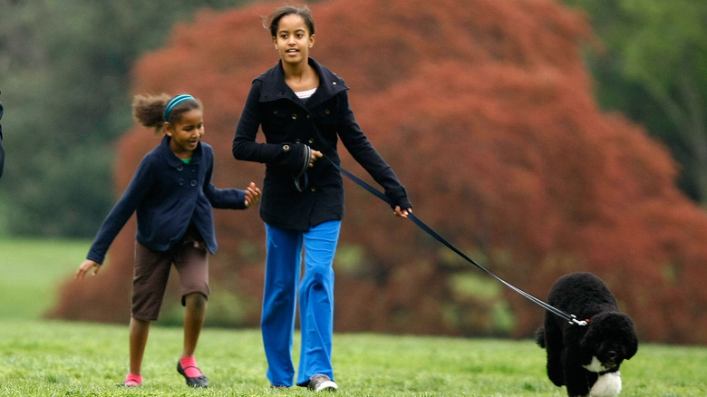 Sasha Obama wearing a blue jacket and brown shorts with Malia Obama wearing a black coat, blue jeans, and holding a dog on a leash