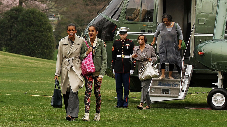 Michelle Obama wearing a tan coat and grey slacks with Malia Obama wearing a green jacket and floral leggings