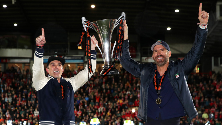 Rob McElhenny and Ryan Reynolds wearing Wrexham AFC gear while holding a trophy