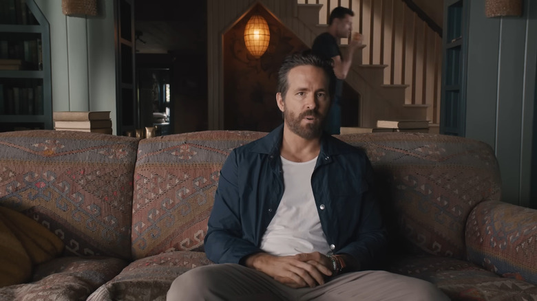 Ryan Reynolds wearing a blue collared shirt and white undershirt on couch with Hugh Jackman in the background