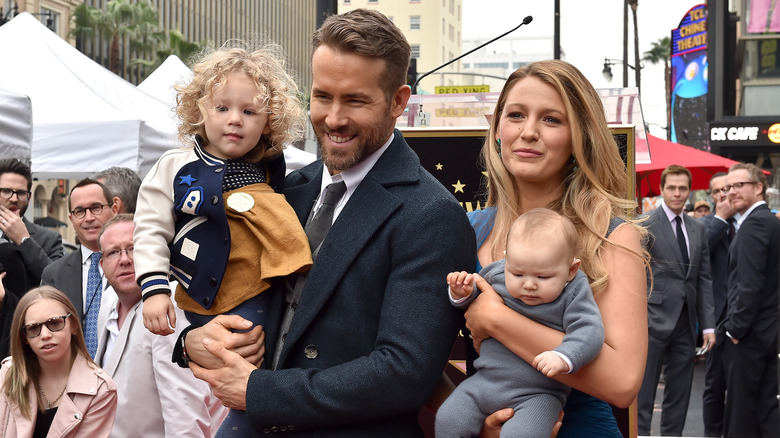 Ryan Reynolds wearing a black suit and tie with Blake Lively in a blue dress while both hold their children