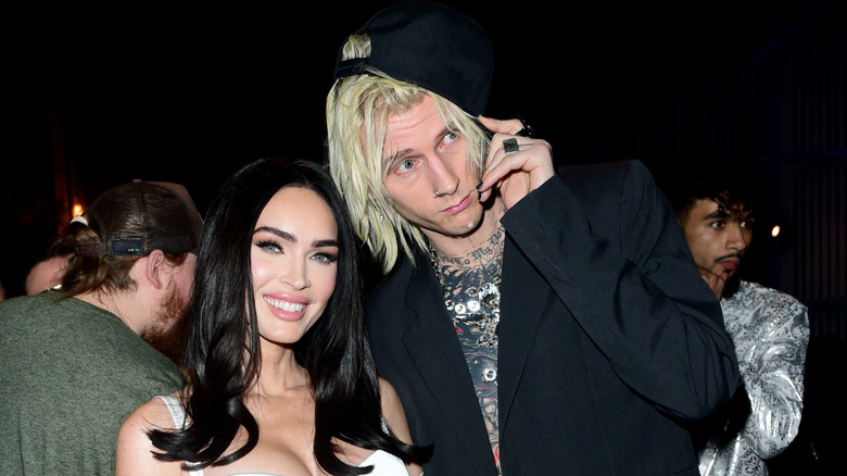 Machine Gun Kelly grabbing brim of his black baseball hat and leaning toward Megan Fox