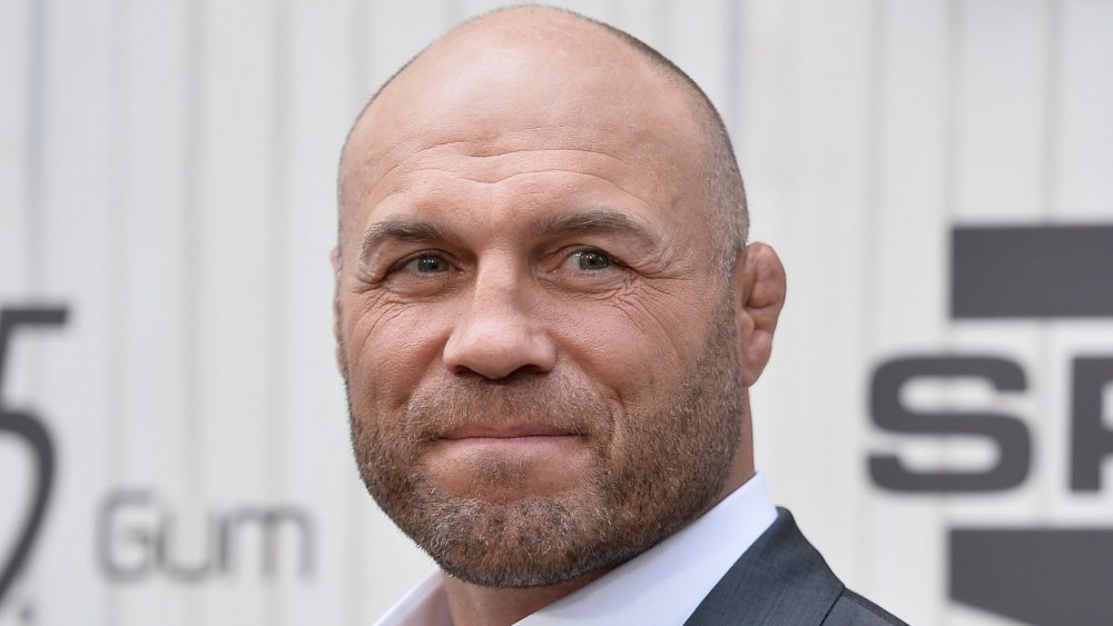 Randy Couture in a dark gray suit and white shirt, looking off to the side with a small smile