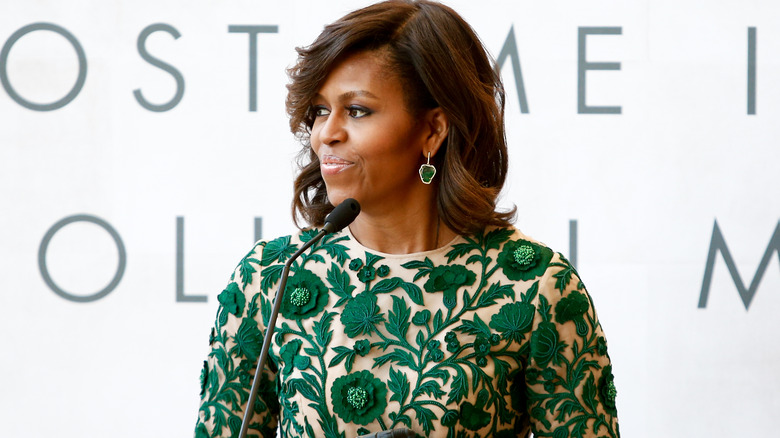 Michelle Obama speaking at an event, wearing green floral dress and matching earrings