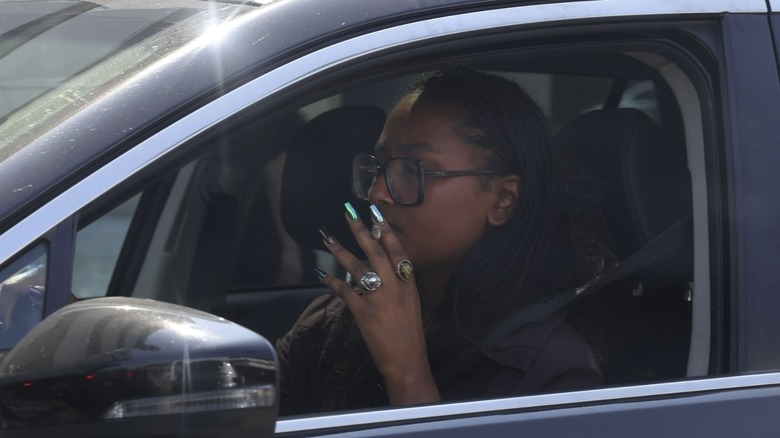 Sasha Obama is seen walking to her car before driving away, enjoying a smoke on the road. Dressed in a dark button-down shirt tucked into light blue high-waisted denim shorts, she completes the look with green slip-on shoes, a large brown handbag, and a bold round necklace. Shot on 09/13/24