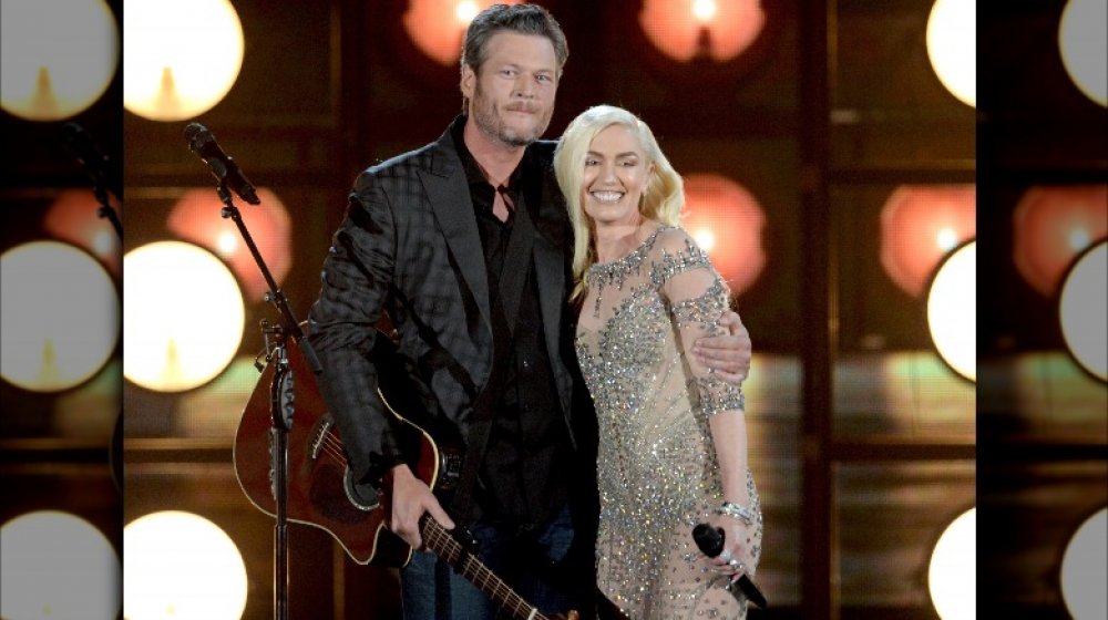 Blake Shelton in a gray-and-black blazer with jeans, Gwen Stefani in a sparkly dress, both smiling on stage