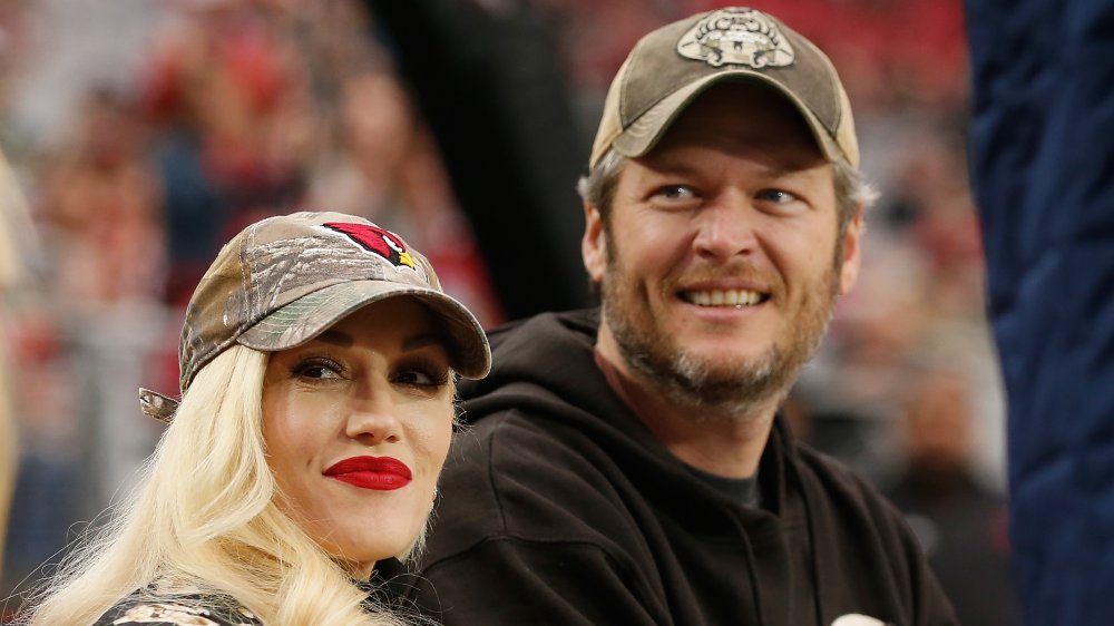 Gwen Stefani and Blake Shelton, both wearing camo baseball hats and looking off to the side