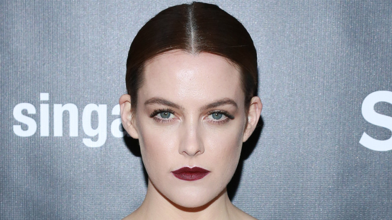 Riley Keough black hair slick, dark lipstick, looking serious