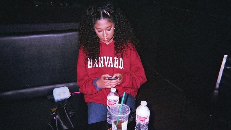 Aoki Lee Simmons on her phone wearing a Harvard hoodie