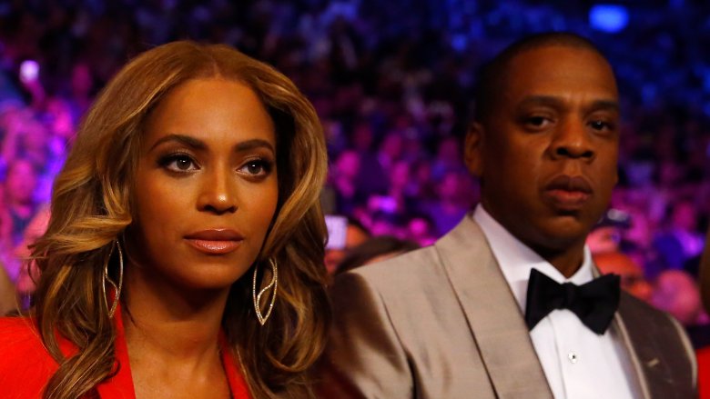 Beyoncé in a red dress, Jay-Z in a silver suit, both looking serious at an event