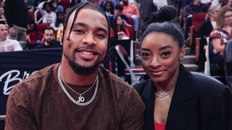 Jonathan Owens wearing a brown shirt and silver necklaces with Simone Biles wearing a black jacket, red undershirt, and silver necklace