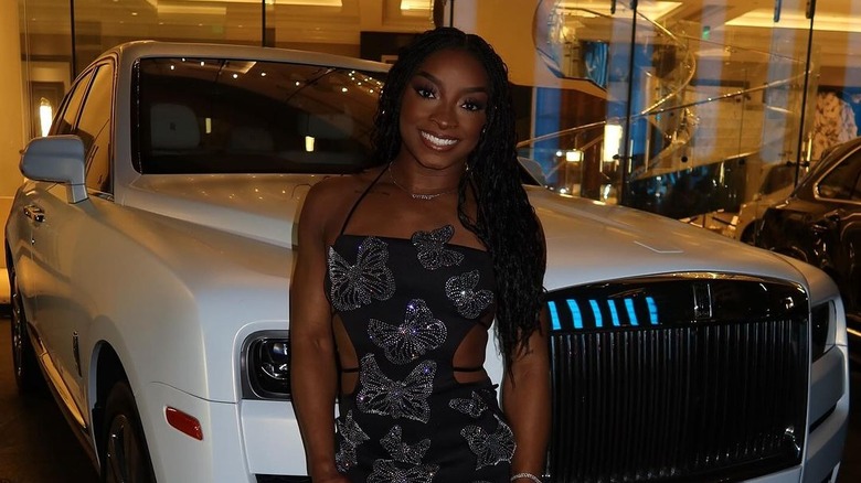 Simone Biles wearing a black embellished dress standing in front of a white SUV