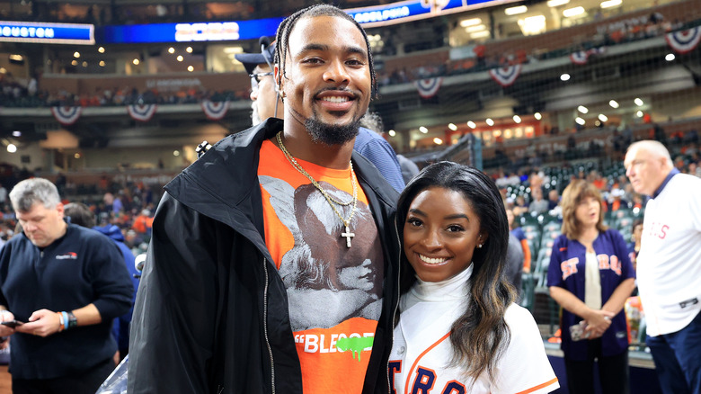 Jonathan Owens wearing an orange graphic t-shirt and black jacket with Simone Biles wearing a white Houston Astros baseball jersey and white turtleneck