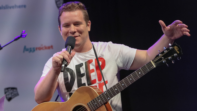 Jon Peter Lewis on stage with a microphone and guitar