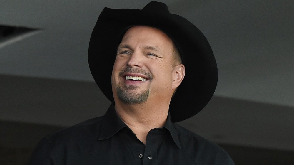 Garth Brooks in a black button-up shirt and cowboy hat, smiling while looking off to the side