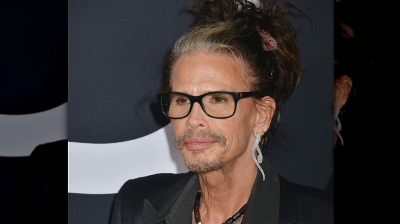 Steven Tyler wearing spectacles on red carpet in a black suit with hair in bun