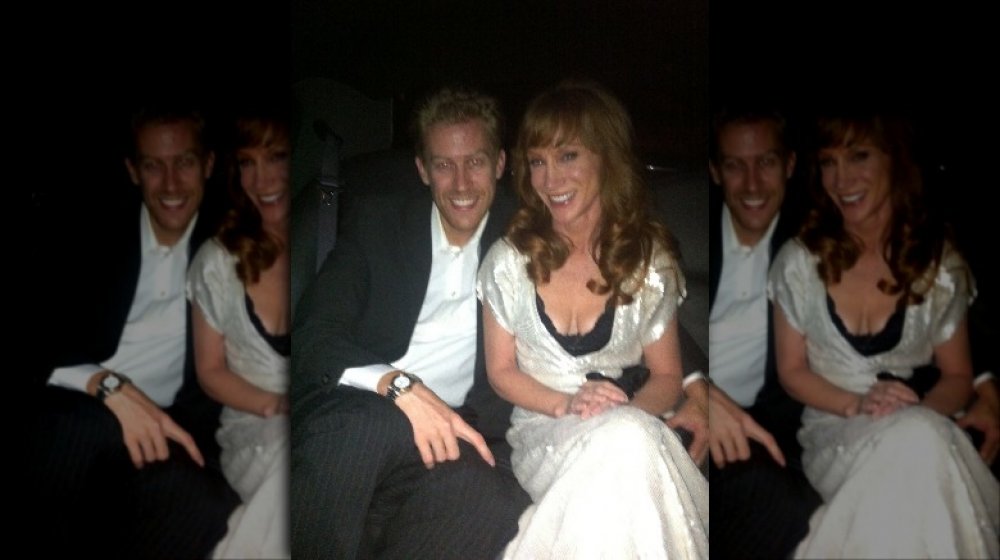 Randy Bick in a black suit, Kathy Griffin in a white dress, sitting in a car and smiling together