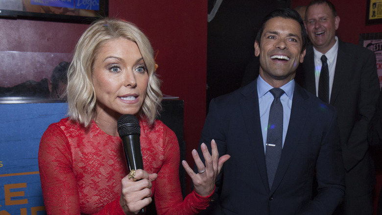 Kelly Ripa talking into a microphone and Mark Consuelos reacting