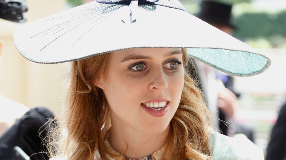 Princess Beatrice in a white outfit and large hat, looking off to the side while smiling