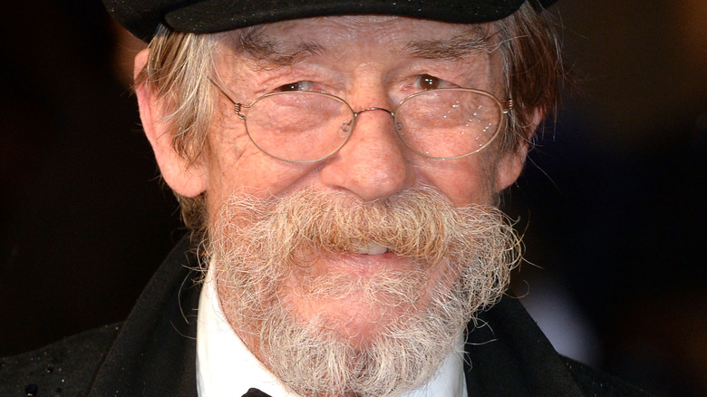 John Hurt smiling under a black cap, wearing glasses