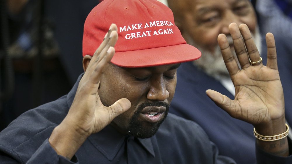 Kanye West with his arms up and eyes closed, wearing a red MAGA hat