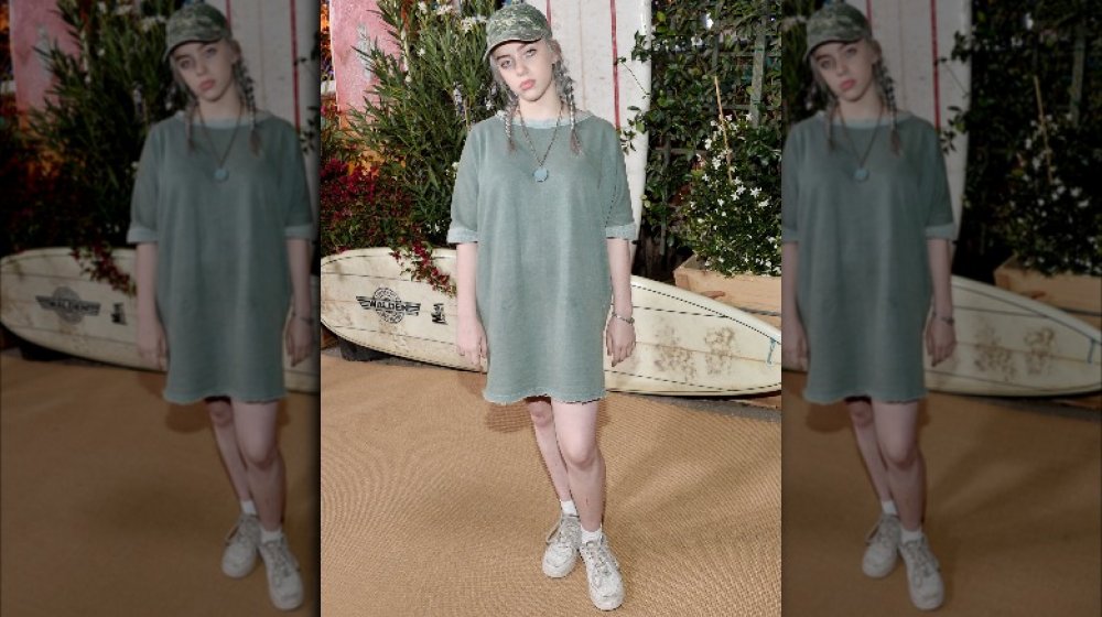 Billie Eilish posing with a solemn expression in a green t-shirt dress, white sneakers, and a camo hat