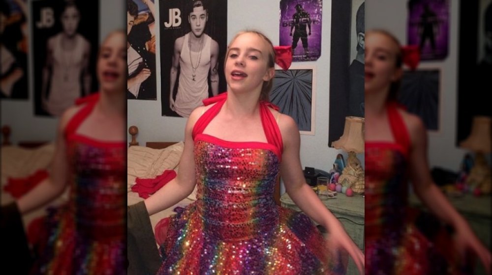 An early teenage Billie Eilish in a sparkly rainbow dress and red bow, standing in front of Justin Bieber posters in her childhood bedroom