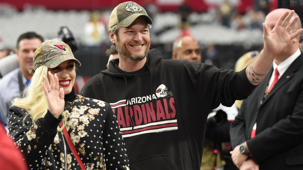 Gwen Stefani and Blake Shelton smiling, wearing baseball caps 