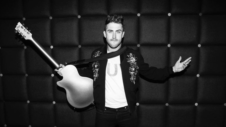 Black and white photo of Jordan McGraw holding a guitar with his arms spread wearing a floral embellished jacket