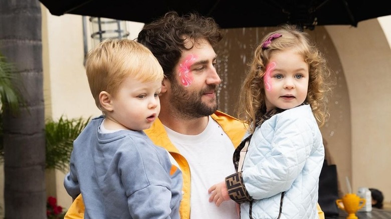 Jordan McGraw holding his two young children on his hips while wearing pink face paint and a yellow jacket