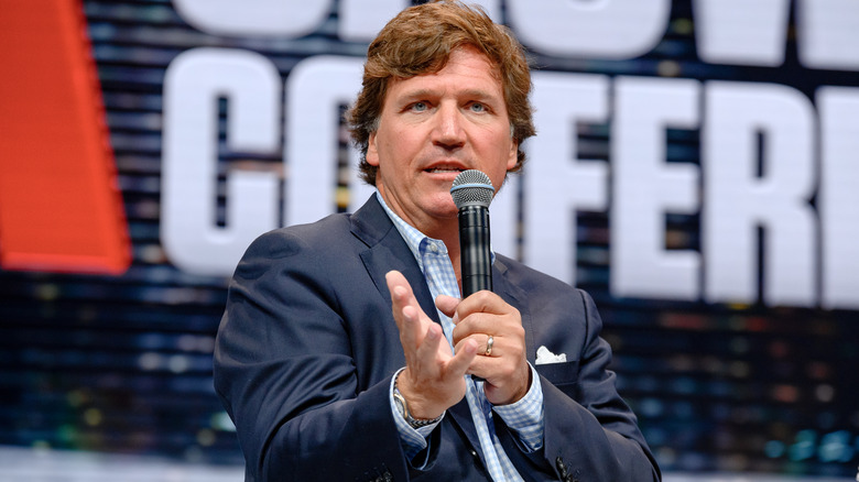 Tucker Carlson holding a microphone while seated on a panel