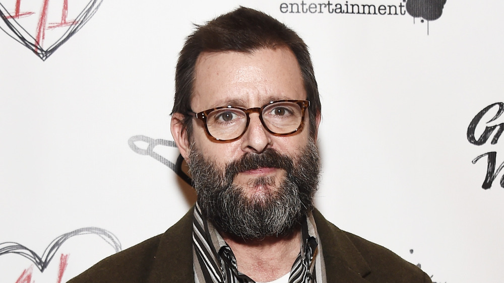 A bearded Judd Nelson smirking, wearing glasses