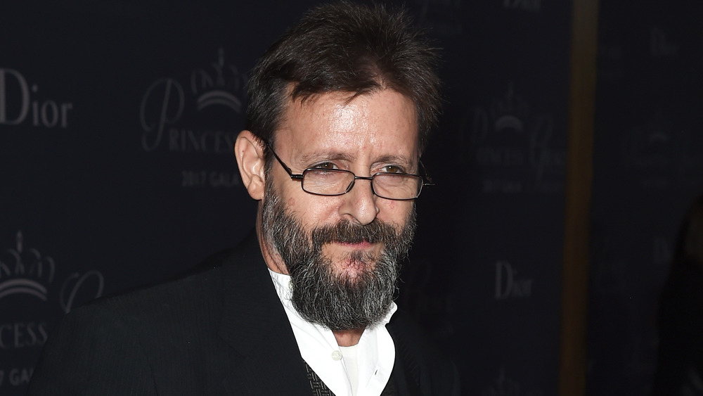 A bearded Judd Nelson smirking and wearing glasses 