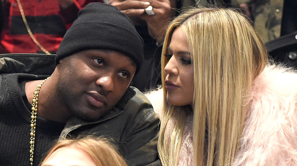 Lamar Odom, wearing a hat, sitting at a fashion show in 2016, leaning towards Khloe; Khloe Kardashian, talking to Lamar Odom, at fashion show in 2016, long blond hair