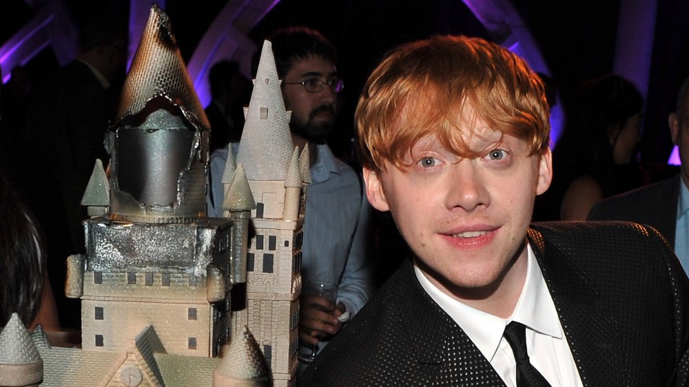 Rupert Grint in a black suit, posing in front of a Hogwarts miniature