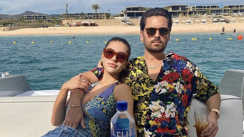 Amelia Hamlin and Scott Disick on a boat, sitting at a table, wearing sunglasses, arms around each other