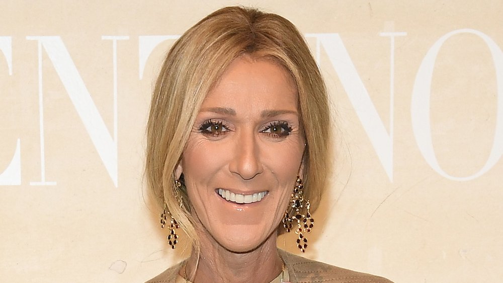 Céline Dion in a beige outfit and dangling earrings, smiling big